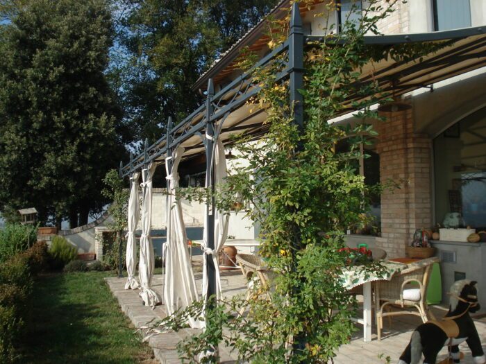Unosider Wandpergola Novecento mit weissen Vorhängen in einem Garten.