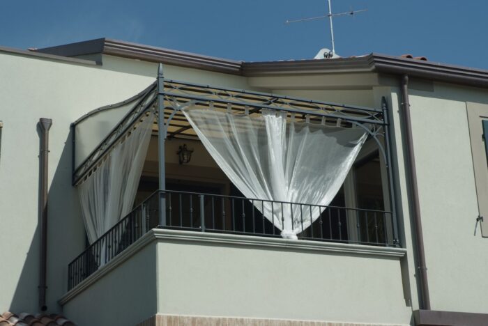 Unodisder Wandpergola Novecento auf einem Balkon mit durchsichtigen, zusammengebundenen Vorhängen