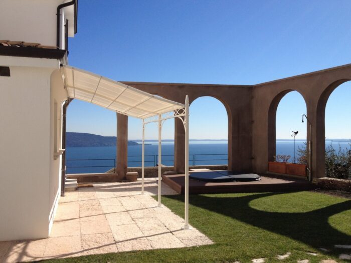 Unosider Wandpergola Novecento in weiss an einem weissen Haus befestigt in einem Garten mit Blick aufs Meer.